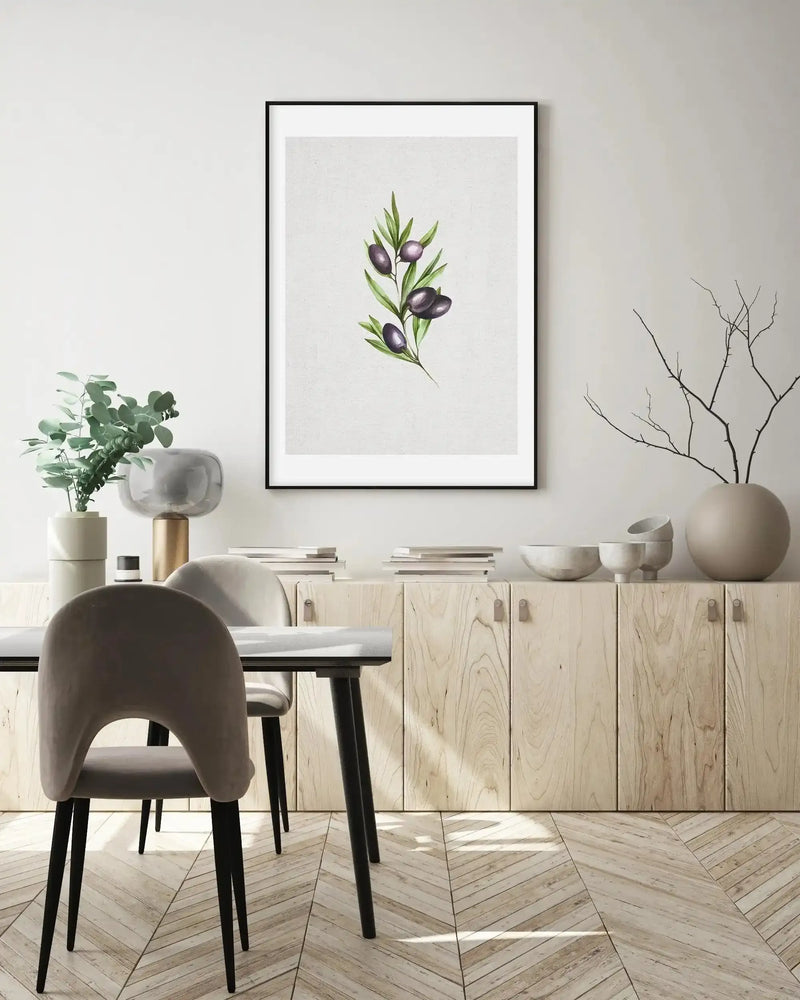 Lifestyle shot of a dining room with a framed art print displayed on a white wall above a light wood cabinet. The print features a watercolour illustration of an olive branch with dark purple olives and green leaves on a textured cream background, with a black frame and white border. The room includes a dining table with two grey velvet chairs, a potted plant, a glass lamp, and decorative bowls.