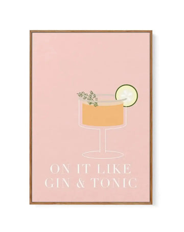 Framed canvas art print with a wooden tan frame featuring an illustrated cocktail on a blush pink background. The drink is rendered in a minimalist style with a clear coupe glass containing golden-amber liquid, garnished with a thin green lime wheel and herb sprigs. Below the illustration, white serif text reads "ON IT LIKE GIN & TONIC". The canvas has a white border around the artwork edges.