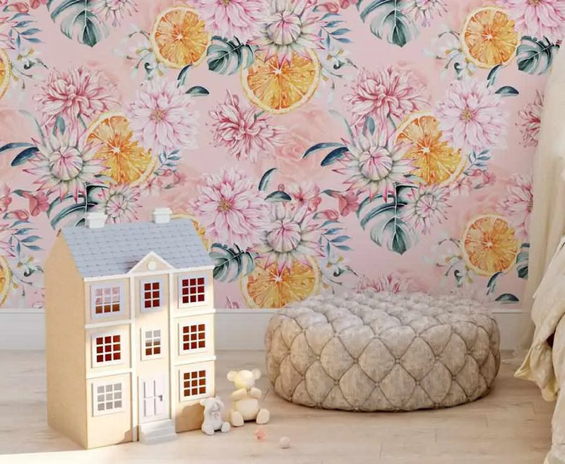Peel and stick wallpaper featuring a repeating pattern of large-scale dahlias and citrus slices in soft pink, white, and orange tones with green leaves on a light pink background, displayed on a wall in a child's bedroom. A light brown dollhouse with white trim and a grey roof sits on the light wood floor, next to a quilted grey ottoman and two small teddy bears.