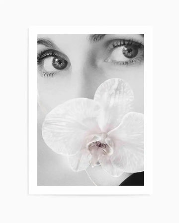 Black and white photography art print of a woman's face with a white orchid covering her mouth, printed on fine art paper and displayed with a white border. The woman's eyes are looking to the side, with long eyelashes and dark eyebrows. The orchid has delicate petals and a subtle pinkish-purple center.