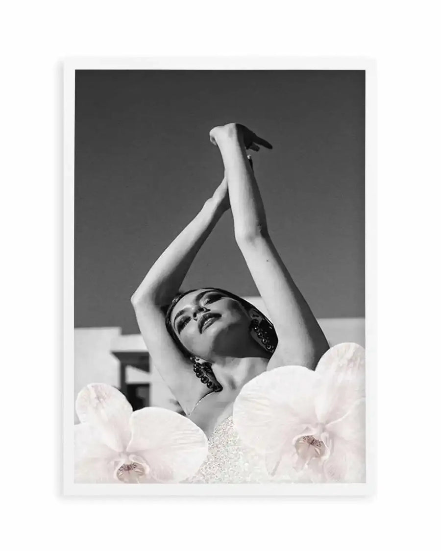 Framed black and white photograph of a woman with her arms raised above her head, looking upwards with her eyes closed. The bottom of the image is adorned with two large white orchid flowers, partially obscuring her body. The woman wears large, dark earrings and a shimmering top. The background is a clear, bright sky with hints of a building structure.