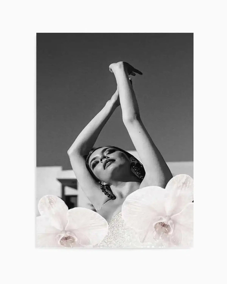 Black and white photography art print featuring a woman with her arms raised above her head, looking upwards with her eyes closed. The composition shows her from the chest up, with two white orchids in the foreground, partially obscuring her body. Her hair is pulled back, and she wears large, dark earrings. The background is a clear sky with a hint of a building structure.