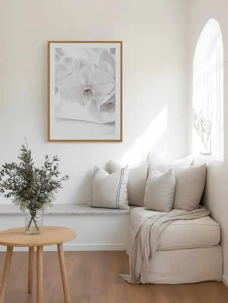 Large framed art print displayed above a built-in corner bench in a bright, minimalist living space. The print features a close-up, soft-focus photograph of white orchid blooms with subtle pinkish-brown centers, printed on fine art paper and displayed in a natural oak frame with white border. The room includes a light cream upholstered bench with matching pillows and throw, a round light wood side table with a vase of green foliage, and an arched window.