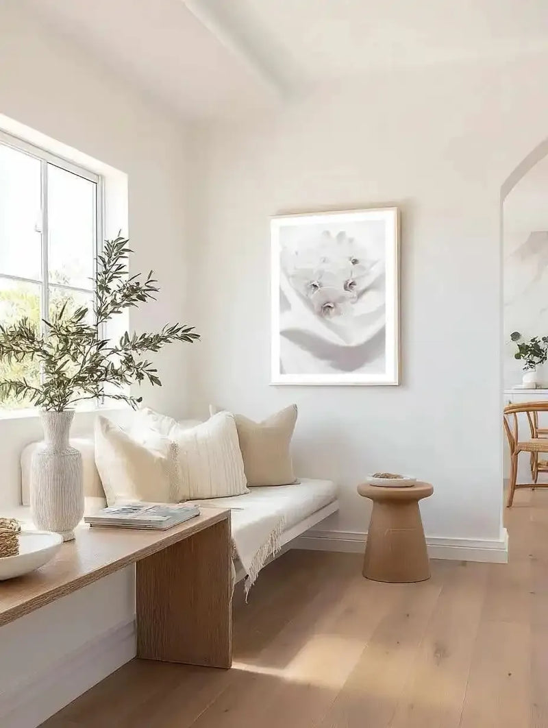 Orchids II Art Print displayed in a natural wood frame with white border, hanging on a cream wall above a built-in bench. The black and white photograph shows delicate orchid flowers in soft focus with fine detail of stamens and petals. The print is shown in a minimalist, light-filled room with cream upholstery, natural wood furniture, and potted greenery, demonstrating the artwork in a serene, modern interior setting.