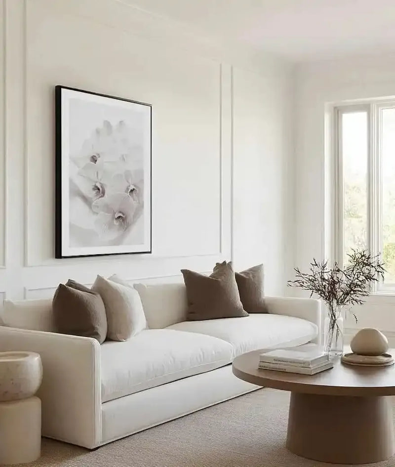 Large framed art print displayed above a white sofa in a minimalist living room. The print is a black and white photograph of three delicate orchid blooms with soft, slightly blurred petals, displayed in a black frame with a white border. The room includes a round wooden coffee table with books and a vase of dried branches, and a cream-colored rug.