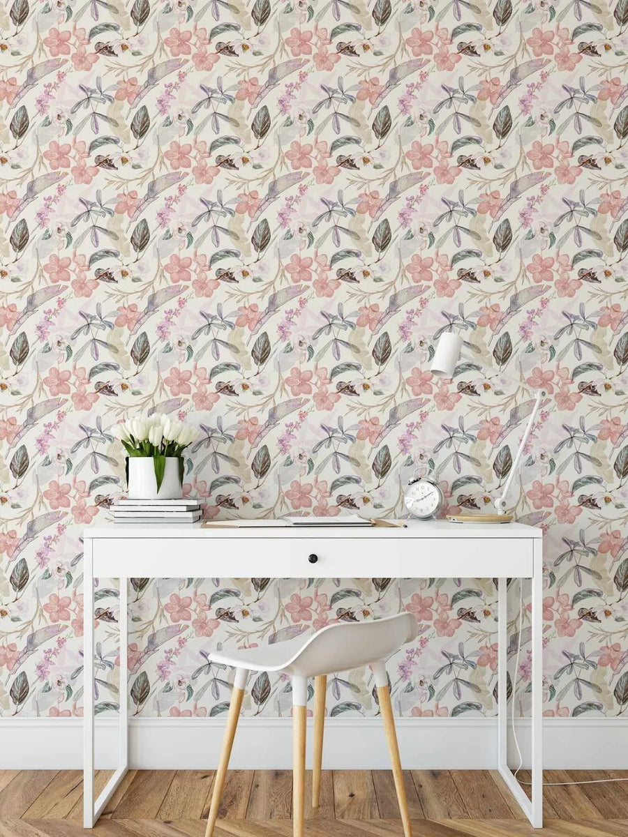 Lifestyle shot of a home office with Oriental Orchids Wallpaper. The wallpaper features a repeating pattern of hand-painted oriental orchids and tropical leaves in soft pink, purple, and muted green tones on a light cream background. The design has a delicate watercolour quality with visible brushstroke texture. A white desk with a drawer, a white chair with wooden legs, a vase of white tulips, books, an alarm clock, and a desk lamp are visible in the foreground.