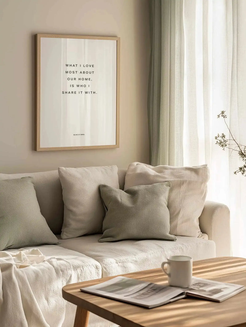 Framed art print displayed on a pale beige wall above a cream linen sofa. The print features black serif text on a cream textured background reading "WHAT I LOVE MOST ABOUT OUR HOME, IS WHO I SHARE IT WITH" with small attribution text at bottom. Natural wood frame with white border. Sheer cream curtains visible to the right, dried botanicals in a clear vase on windowsill, cream throw pillows and blanket on sofa, wooden table with magazines and ceramic mug in foreground.