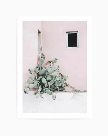 Photography art print featuring a prickly pear cactus with pale green pads and small red fruits growing against a pastel pink wall with a small, dark rectangular window and a narrow vertical opening. The cactus sits atop a white ledge, with some tendrils trailing along the wall.