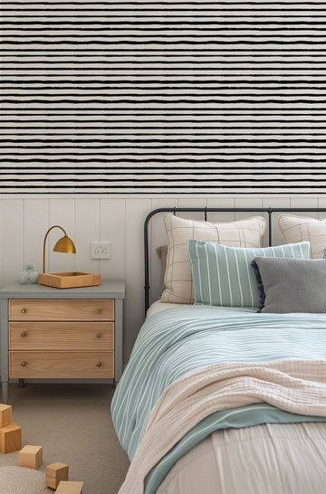 Lifestyle shot of a bedroom featuring Painterly Horizontal Stripe Wallpaper above a white wainscoting wall. The wallpaper has hand-painted horizontal stripes in black on a light cream background, with a loose, organic brushstroke quality. The room includes a bed with a black metal frame, light blue and white striped duvet cover, and various pillows in cream, light blue, and grey. A grey bedside table with wooden drawers and a brass lamp is visible on the left.