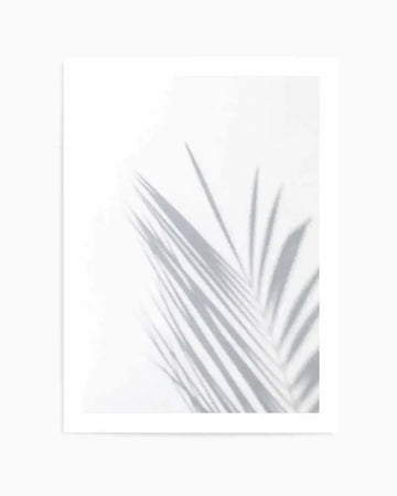 Minimalist art print featuring a single palm frond with elongated, feathered leaflets in soft gray tones against a pale cream background. The frond is positioned in the lower right portion of the composition, creating negative space in the upper left. The image has a soft, slightly blurred quality suggesting botanical photography or watercolor illustration. Displayed in a thin white frame with white border.