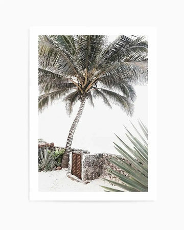 Framed black and white photograph of a tropical resort scene with a natural wood frame and white border. The composition features a tall palm tree with a curved trunk on a sandy beach, with a low stone wall and a wooden gate in the foreground, and large agave-like plants on the right.