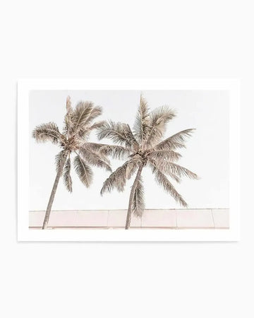Framed photograph of two palm trees against a bright white sky, with a light pink-beige wall visible behind their trunks. The palm fronds are a muted, desaturated brown-grey, giving the image a soft, minimalist aesthetic.
