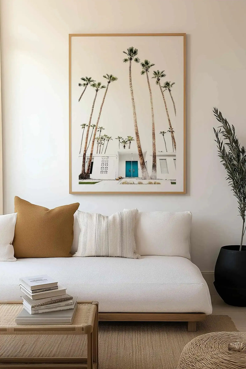 Framed art print featuring a soft-focus photograph of tall palm trees with textured trunks against a white modernist building with a turquoise door, displayed in a natural wood frame with white border above a cream linen sofa styled with tan, striped, and cream pillows in a minimalist living room.
