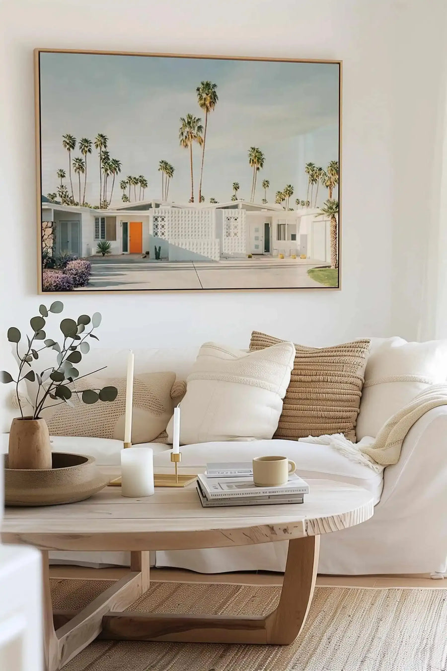 Framed canvas art print depicting a mid-century modern house with white exterior and orange door, surrounded by tall palm trees against a pale blue sky. The single-story residence features geometric white lattice panels and manicured landscaping with purple flowering plants. Displayed above a cream linen sofa in a minimalist living room styled with natural wood furniture, woven textiles, eucalyptus stems in a wooden vase, and a light wood coffee table. The print has a natural wood frame with white border.