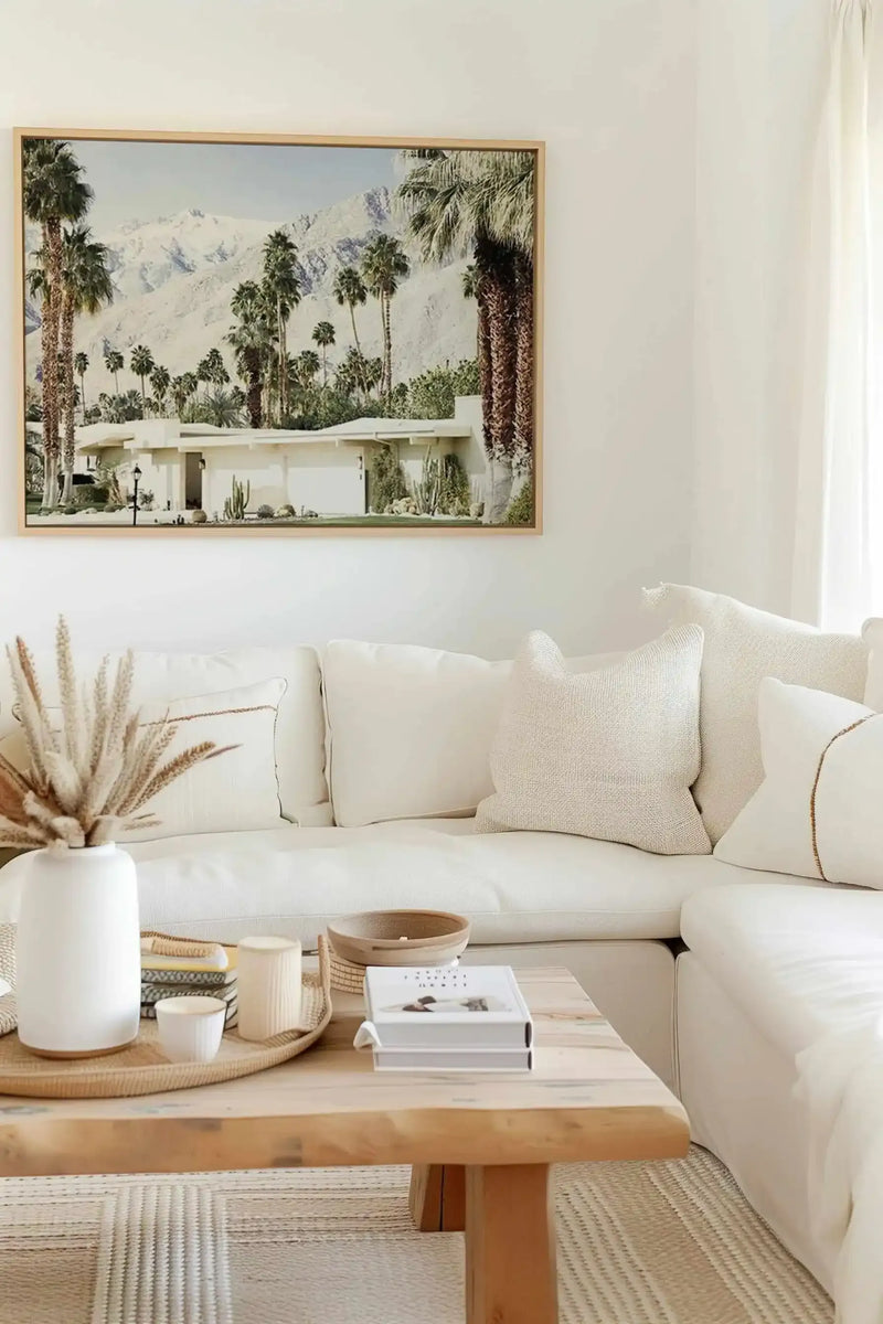 Framed canvas art print depicting a mid-century modern white house nestled against snow-capped mountains, surrounded by tall palm trees in various shades of green. The photograph features a clear blue sky and manicured desert landscaping. The print is displayed in a natural wood frame with white border, hung on a cream-colored wall above a cream linen sofa styled with textured throw pillows and a wooden coffee table decorated with a white ceramic vase, woven baskets, and stacked books.