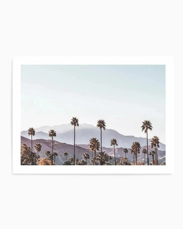 Product shot of a framed photography art print featuring a desert landscape with tall palm trees in the foreground and layered mountains in the background under a pale blue sky. The scene has a warm, hazy quality with soft light.