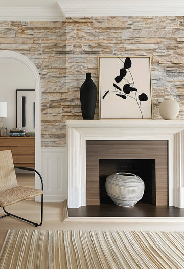 "Palm Springs Sandstone Wall Wallpaper applied to a living room wall, featuring a stacked stone pattern in varying shades of beige, cream, and light brown, creating a textured, rustic look. The wallpaper is visible behind a white fireplace mantel decorated with a black vase, a framed abstract art print with black organic shapes on a cream background, and a cream-coloured vase. To the left, an arched doorway leads to another room with a wooden dresser and a woven chair."