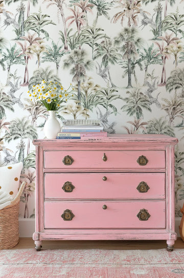 "Tropical wallpaper featuring a repeating pattern of hand-painted palm trees and cranes in muted sage green, dusty olive, and warm brown tones on a soft white background. The botanical design has a loose, organic watercolour quality with visible brushstroke texture. The wallpaper is displayed behind a distressed pink dresser with antique brass hardware, a white vase with daisies, and a stack of books."