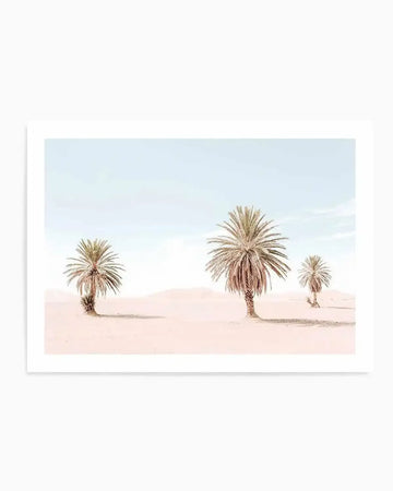 Photography art print featuring three palm trees in a desert landscape under a pale blue sky. The sand is a soft, light pinkish-beige, and the palm trees have light brown trunks and dried, light green to beige fronds. The composition shows the largest palm tree in the mid-ground to the right, a smaller one to the left, and a very small one further back on the right, with gentle sand dunes in the background.