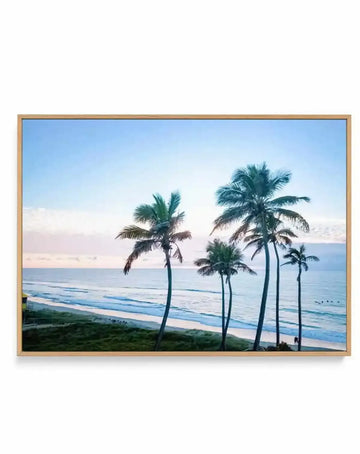 Framed canvas art print featuring a serene beach landscape with tall palm trees silhouetted against a soft pastel sky. The scene shows calm ocean waves in shades of blue and turquoise, sandy beach in the foreground, and a dreamy sunrise or sunset with pink and lavender tones on the horizon. The artwork is displayed in a natural wood frame with a white border, creating a coastal, tranquil atmosphere.