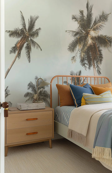 Peel and stick wallpaper photo mural featuring a tropical scene with large palm trees against a light blue, slightly overcast sky, displayed in a bedroom. The bed has a light wood headboard, blue and white striped sheets, and an assortment of pillows in shades of blue, mustard yellow, and textured rust orange. A light wood nightstand with leather pull handles and a small green lamp sits beside the bed.