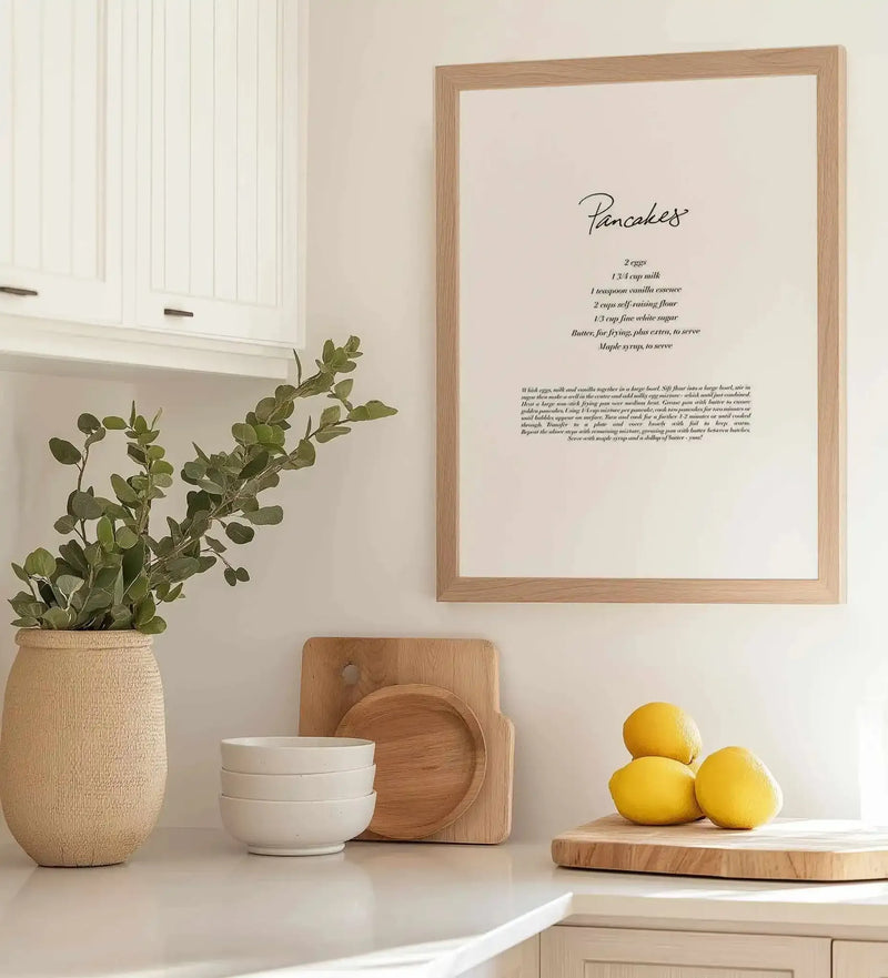 Framed art print displayed on a cream wall showing "Pancakes" handwritten in script at the top, followed by a recipe ingredient list and cooking instructions in black italic text on a cream background. Natural wood frame with white border. Styled on a kitchen counter with a woven tan vase containing eucalyptus branches, white ceramic bowls, wooden cutting board and circular wooden trivet, and three yellow lemons on a wooden tray.