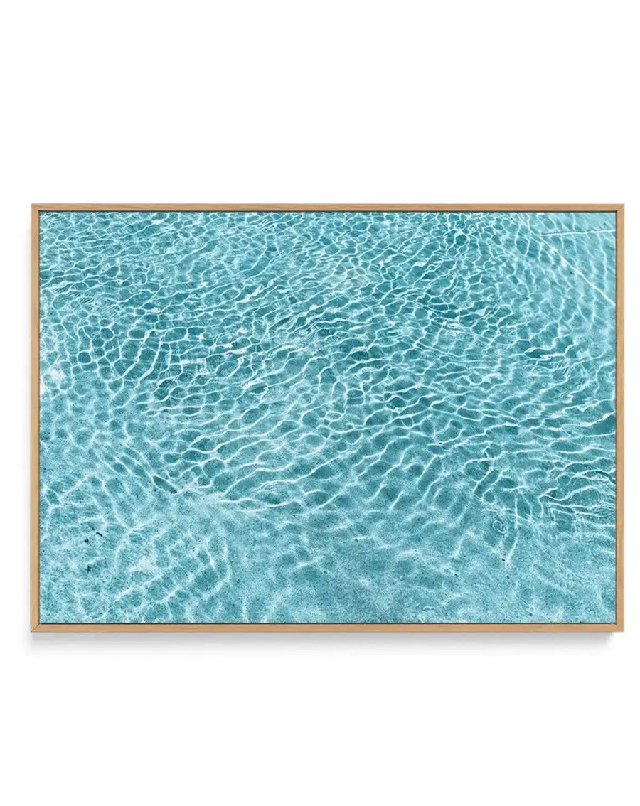 Framed canvas art print titled Paradise featuring an aerial photograph of shallow turquoise water with rippling wave patterns and sunlight reflections creating intricate textures across the surface. The image transitions from lighter aquamarine tones at the top to deeper teal-blue hues toward the bottom, with white sandy bottom visible through the clear water. Natural wood frame with subtle grain detail surrounds the canvas print.