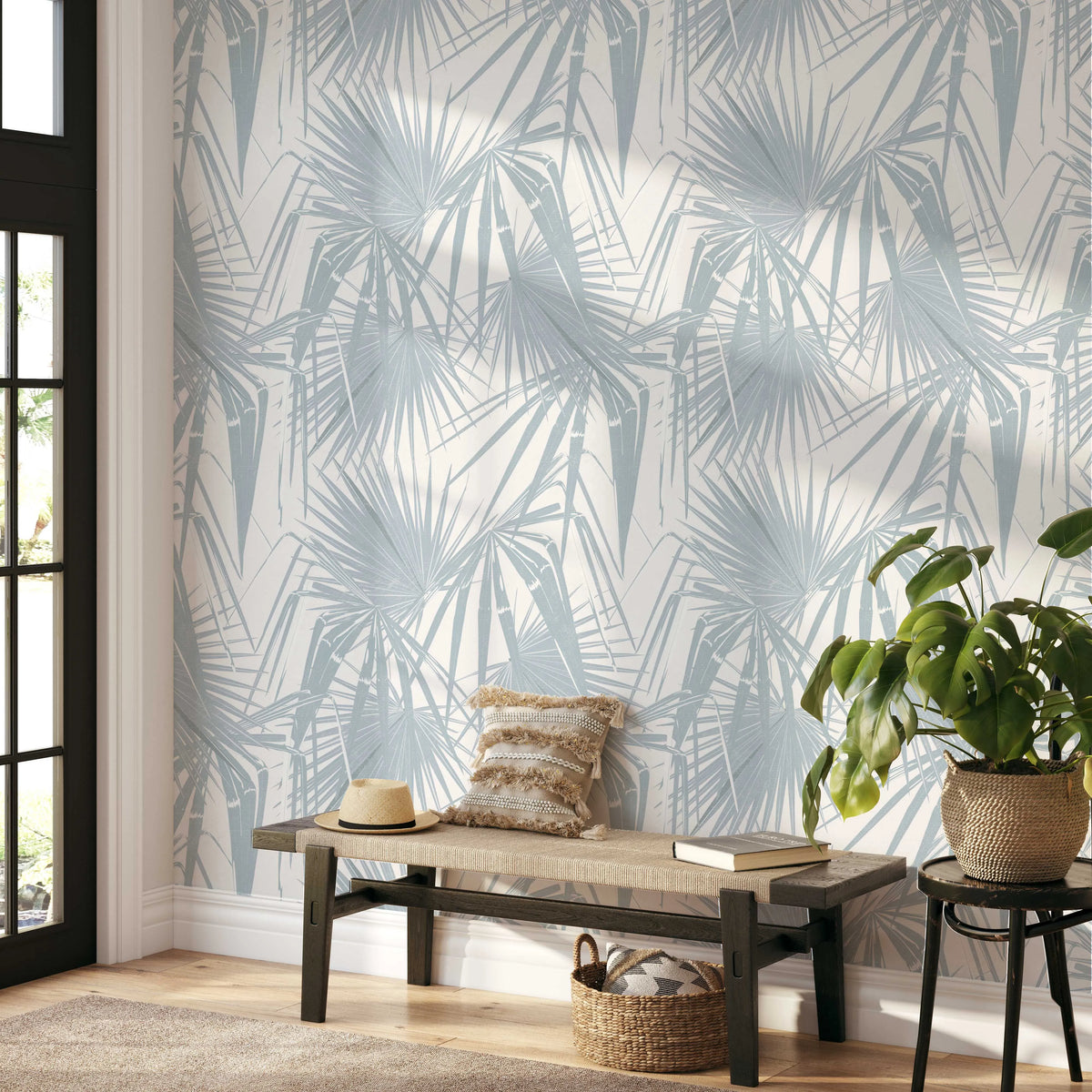 Lifestyle shot of an entryway featuring Paradiso in Ocean wallpaper. The wallpaper has a repeating pattern of large, overlapping palm fronds in a muted, dusty blue-grey against a warm off-white background. Sunlight casts diagonal shadows across the wall. A dark wood bench with a woven beige seat holds a textured cream and brown pillow and a straw hat. A woven basket with a patterned pillow sits beneath the bench. To the right, a dark wood stool holds a potted Monstera plant with large, glossy green leaves.