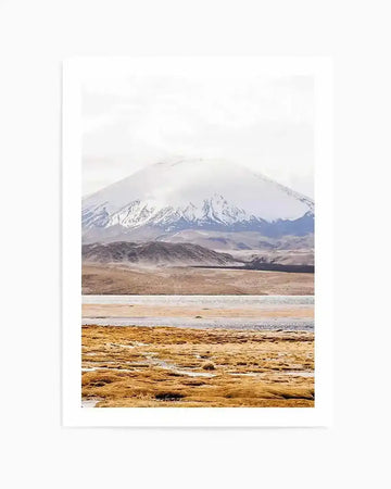 Photography art print featuring a vast, serene landscape with a snow-capped mountain under a bright, overcast sky. The composition shows a large, white mountain peak in the distance, partially obscured by clouds, with rugged, snow-dusted lower slopes. In the midground, a wide expanse of dry, light brown and tan land stretches, leading to a calm body of water with reflections. The foreground is dominated by a field of golden-brown, textured grasses, creating a sense of depth and natural beauty.