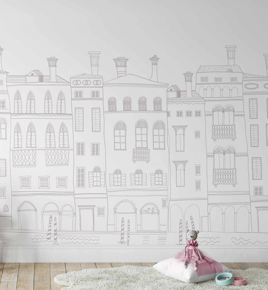 Lifestyle shot of a child's room featuring a peel and stick wallpaper mural with a hand-drawn illustration of a Parisian street scene in light grey on a white background. The mural depicts a row of tall, narrow buildings with various window styles, balconies, and rooftops, some with chimneys, and the suggestion of water with gondola poles at the base. A white shaggy rug is on a light wood floor, with a white pillow and a grey teddy bear in a pink dress sitting on it, next to two pastel-colored wooden rings.