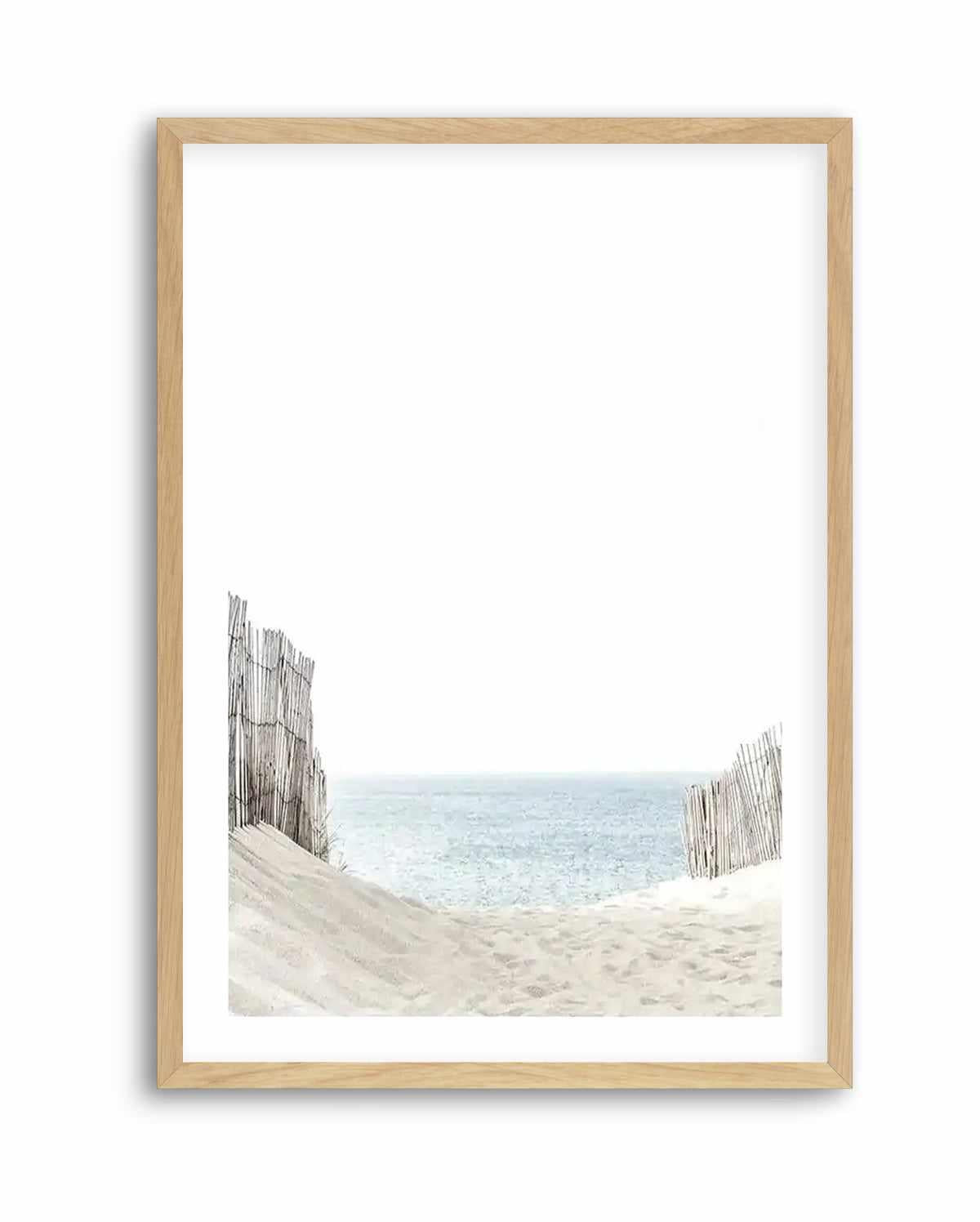 Natural wood frame displaying a serene coastal photograph titled "Path to the Sea Art Print." The artwork features weathered wooden dune fences in gray-brown tones flanking sandy beach in the foreground, with calm turquoise and pale blue ocean water stretching across the middle distance under a clear white sky. The image has a soft, muted color palette with delicate watercolor-like quality. White border surrounds the photograph within the light natural wood frame.