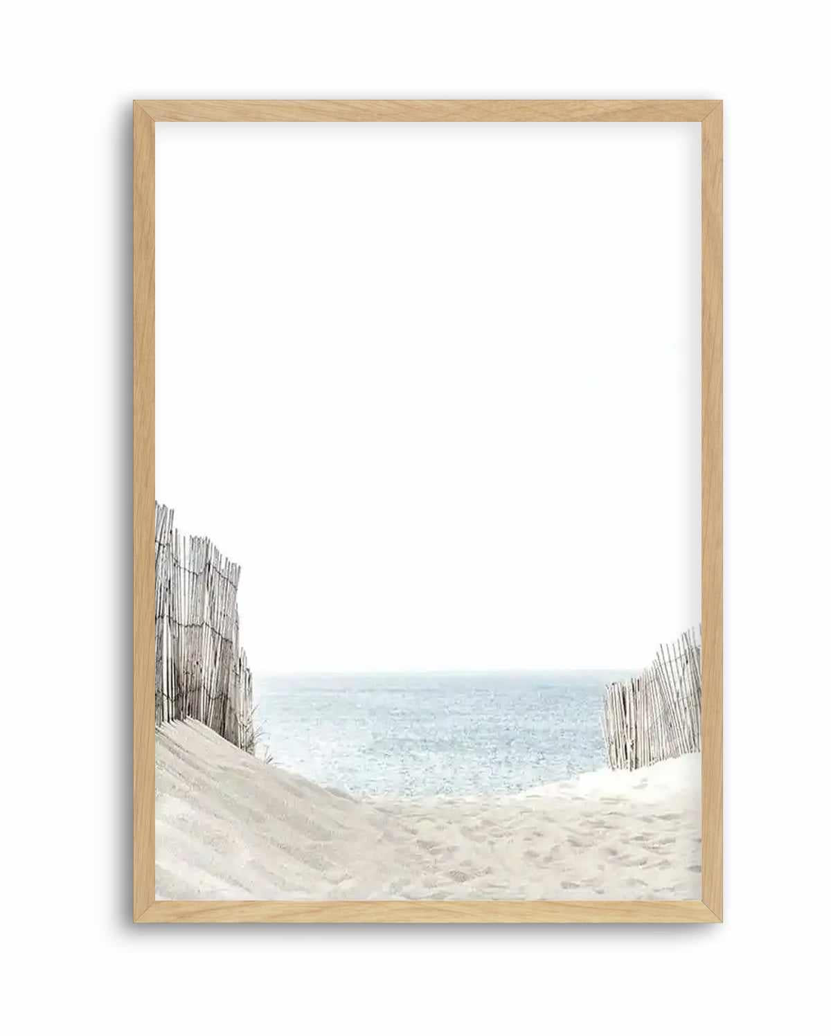Framed art print showing a serene beach scene viewed through weathered wooden fence posts. Sandy dunes in soft beige tones occupy the lower portion, with two gray driftwood fence sections flanking either side. Calm turquoise ocean water stretches across the middle distance under a pale sky. The artwork has a soft, muted color palette with a peaceful, coastal aesthetic. Light natural wood frame with white border.
