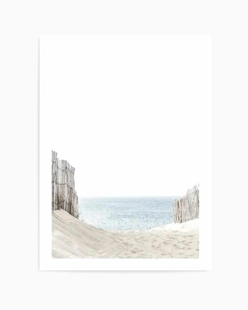 Path to the Sea Art Print displayed on white background. The artwork features a serene beach scene framed by two weathered wooden sand fence sections on either side. Soft, pale beige sand occupies the foreground, leading to calm, pale blue-green ocean waters meeting a hazy horizon. The composition uses muted, naturalistic colors with a soft, dreamy quality suggesting watercolor or photographic imagery. The print is mounted with a clean white border on white paper stock.