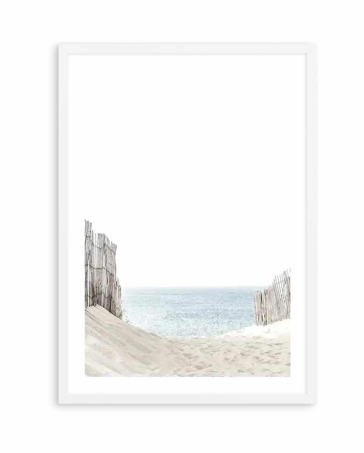 Framed art print featuring a minimalist photograph of a sandy beach scene. Two weathered wooden fence posts frame the composition on either side, with soft beige sand in the foreground and calm turquoise-blue ocean water meeting a pale sky at the horizon. The image has a serene, coastal aesthetic with muted, natural tones. The artwork is displayed in a white wooden frame with a white border.