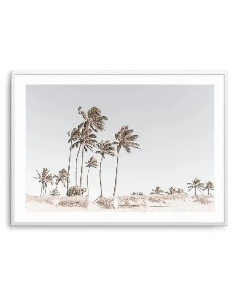 Framed photography art print of a coastal beach scene with a white frame and white border. The composition features numerous palm trees with wind-blown fronds on a sandy dune, under a pale grey sky, creating a serene, minimalist aesthetic.