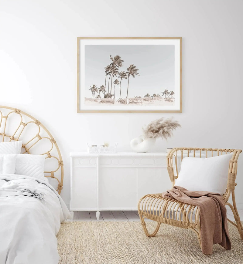 Lifestyle shot of a bright bedroom featuring a large framed art print above a white dresser. The print is a photograph of a sandy beach with numerous palm trees under a pale blue sky, displayed in a natural wood frame with a white border. To the left, a bed with a rattan headboard and white bedding is visible. To the right, a rattan chair with a white pillow and a brown throw blanket sits on a textured natural fiber rug.