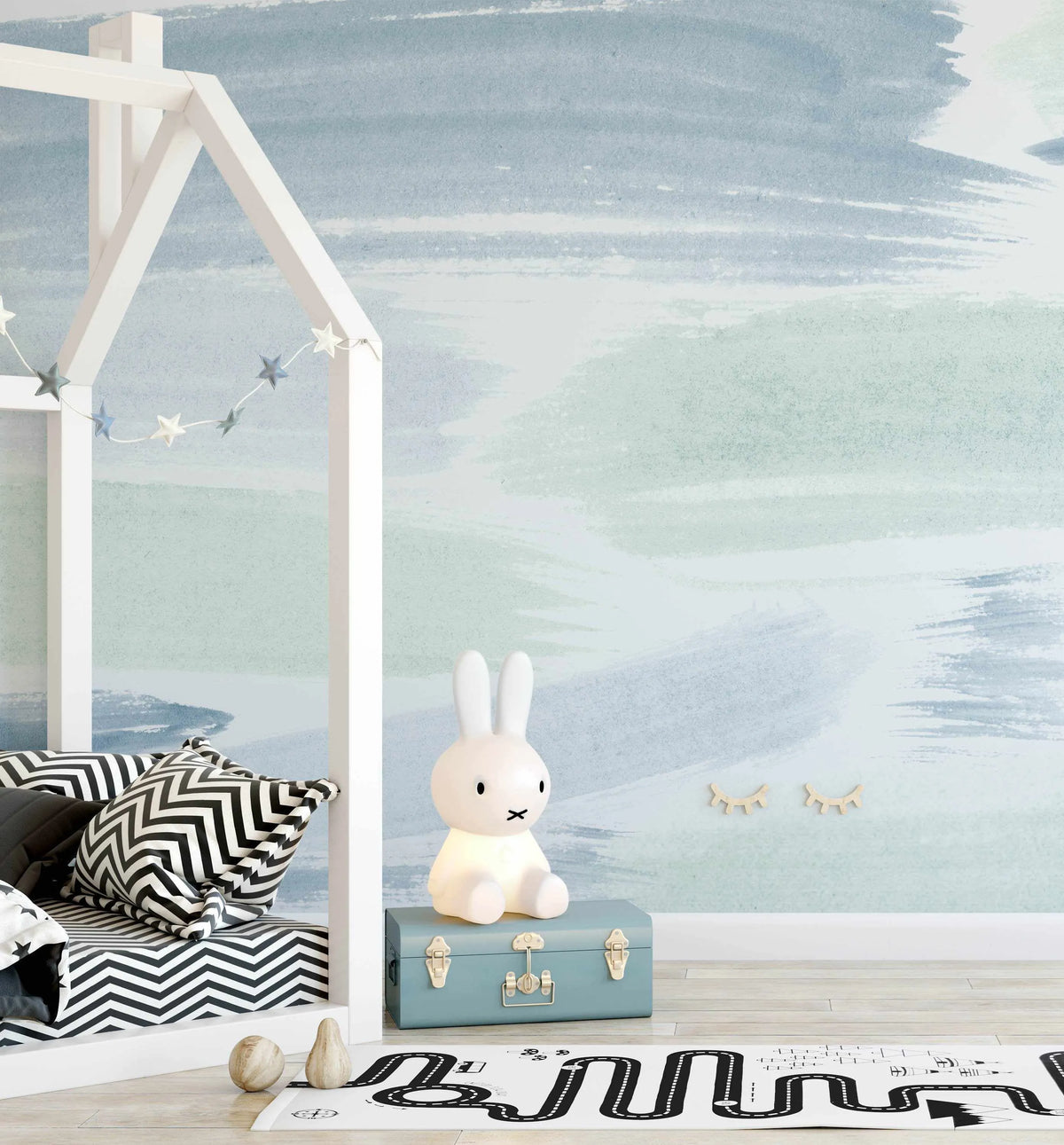 Kids Bedroom:
"Peel and stick wallpaper featuring a repeating abstract pattern of broad, horizontal brushstrokes in muted shades of pale blue, dusty sage green, and soft white, creating a tranquil, watercolour-like effect. The wallpaper is displayed in a child's bedroom with a white house-shaped bed frame, a Miffy bunny lamp on a blue trunk, and a black and white road-themed rug."