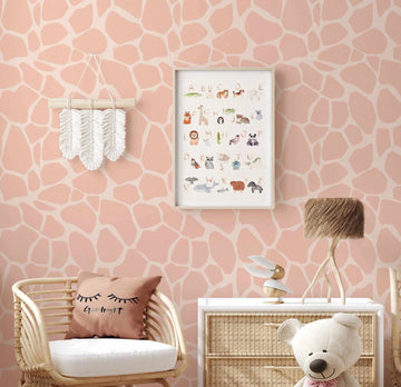 Peachy Giraffe Wallpaper displayed on a nursery wall with an organic giraffe spot pattern in soft blush pink and cream. The wallpaper features irregular stone-like shapes in warm peachy tones with white outlines creating a natural texture. The room shows the wallpaper as backdrop for a woven rattan chair with white cushion, a framed animal alphabet print, decorative macramé wall hanging, woven dresser with white top, and stuffed teddy bear, creating a calm, neutral aesthetic suitable for a child's bedroom.