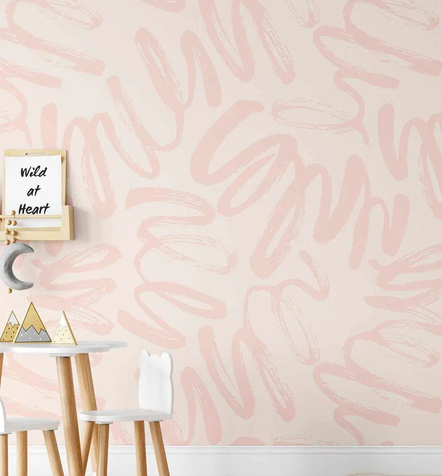 Lifestyle shot of a child's room featuring Peachy Graffiti Wallpaper with a repeating pattern of loose, hand-painted brushstrokes in a soft peach-pink on a light cream background. The wallpaper covers a full wall behind a small white table with two white chairs, a wooden shelf holding a "Wild at Heart" sign, and a grey crescent moon decoration.