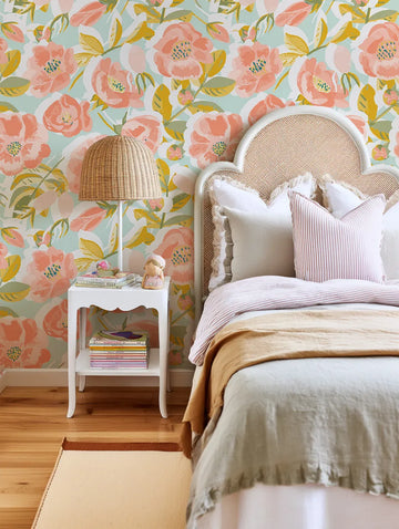 Lifestyle shot of a child's bedroom featuring Peachy Fleur wallpaper behind a bed with a white wicker headboard. The peel and stick wallpaper has a repeating pattern of large-scale hand-painted flowers in shades of peach and coral, with yellow and sage green leaves, on a light blue background. A white scalloped nightstand with a woven lamp and stacked books is to the left of the bed. The bed has white and striped pillows, a light green duvet, and a tan throw blanket.