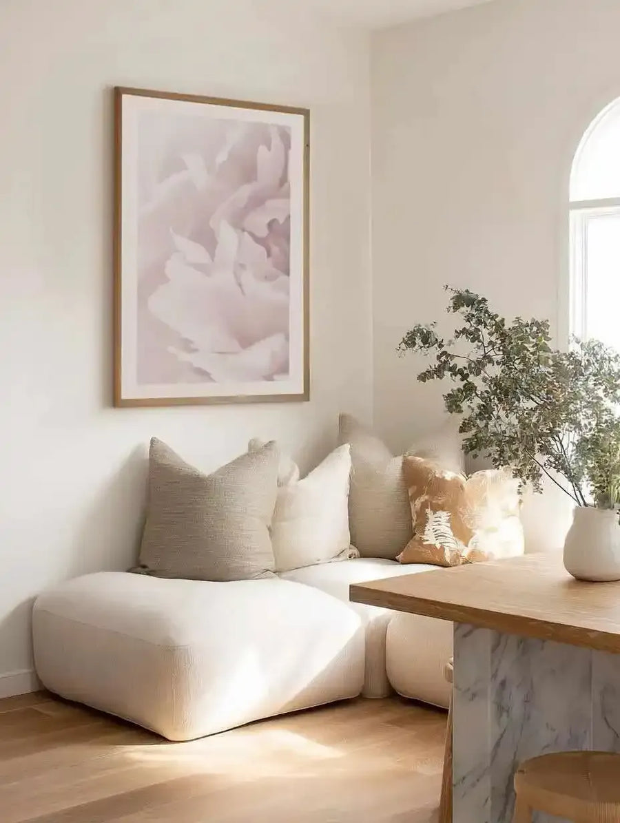 Framed art print displaying soft, abstract peony flowers in pale blush pink and cream tones with a watercolour-like aesthetic. The wooden frame is natural tan/beige with a white border. The print is hung on a cream wall above a curved white upholstered chair styled with neutral linen pillows, positioned next to a light wood table with a potted green plant and white ceramic vase in bright natural light from an arched window.