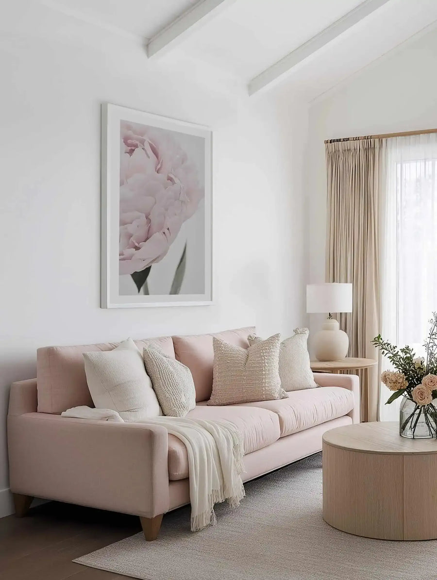 Framed art print of a close-up peony flower with soft blush pink petals and green stem against a pale background, displayed in a white frame above a blush pink sofa styled with cream and neutral patterned pillows in a minimalist living room with natural wood accents and warm lighting.