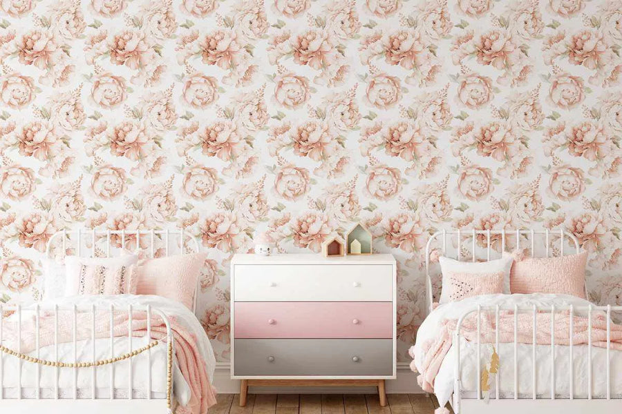 A children's bedroom with two white metal-frame beds flanking a white, pink, and grey three-drawer dresser, all against a wallpaper featuring a repeating pattern of large-scale peony roses in soft terracotta and blush pink with green leaves on a white background. The beds are dressed with white bedding and pink throws and pillows.
