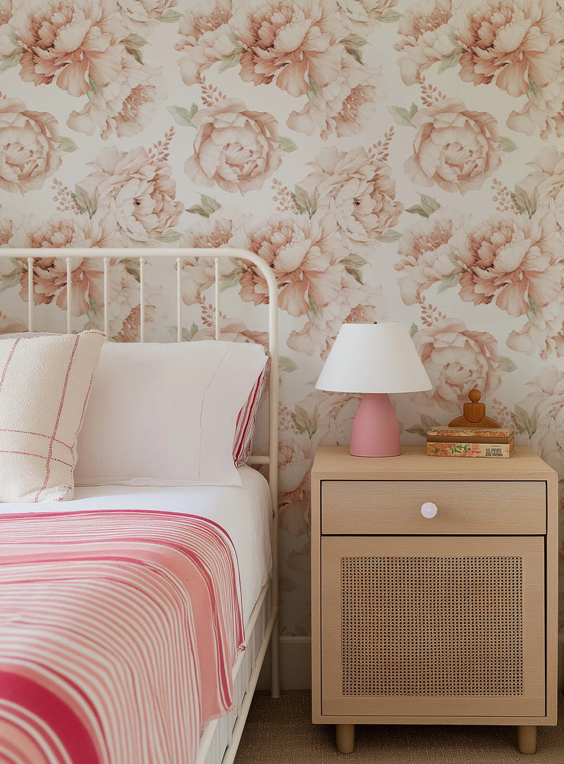Bedroom wall featuring Peony Roses in Soft Terracotta Wallpaper, displaying a repeating pattern of large watercolor peonies in dusty rose and terracotta tones with sage green foliage on a cream background. A white metal bed frame with pink striped bedding and cream pillows sits against the wallpapered wall. A light wood nightstand with woven rattan cabinet door and white ceramic table lamp with pink base is positioned beside the bed.