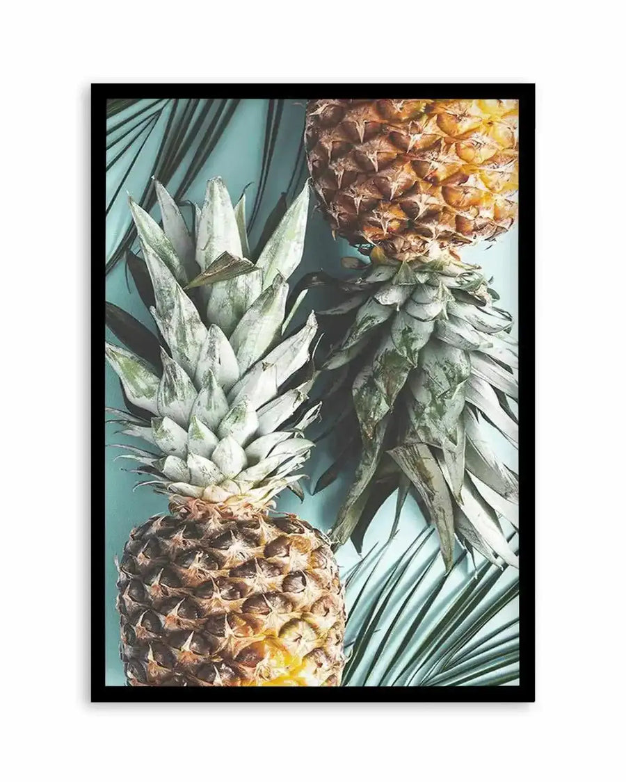 Framed art print featuring a top-down photographic composition of two pineapples with their green leafy crowns, set against a light blue-green background with dark green palm fronds. The pineapples have golden-brown, textured skin and hints of yellow flesh. The print is displayed in a black frame.