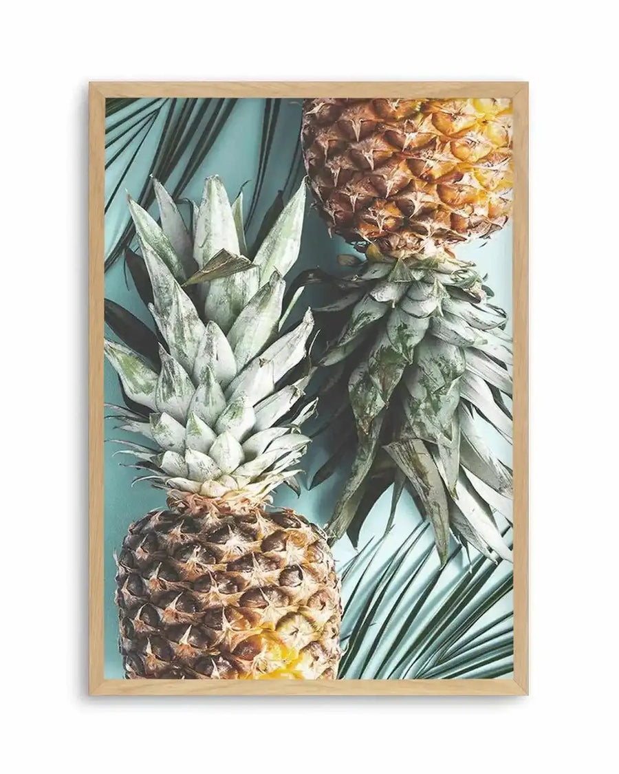 Framed art print featuring a top-down photograph of two pineapples and palm fronds on a light teal background, displayed in a natural wood frame. The composition shows one whole pineapple with its spiky green crown at the bottom left, and another pineapple cut in half horizontally at the top right, revealing its yellow flesh. Dark green palm fronds are scattered across the teal surface.