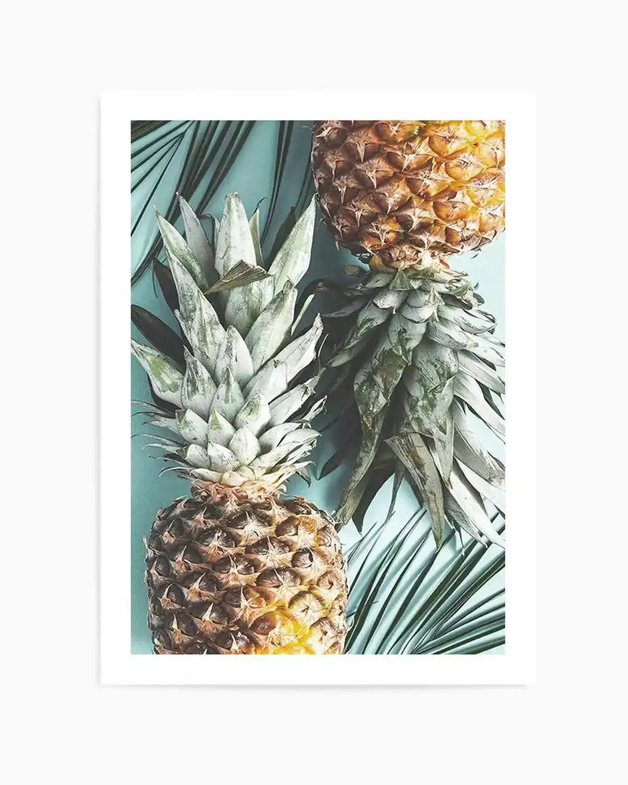 "Photography art print featuring two pineapples and palm fronds on a light blue background. The top pineapple is cut in half horizontally, revealing its golden-yellow flesh, while the bottom pineapple is whole, with its spiky green crown pointing upwards. The palm fronds are a deep emerald green, adding a tropical touch to the composition."