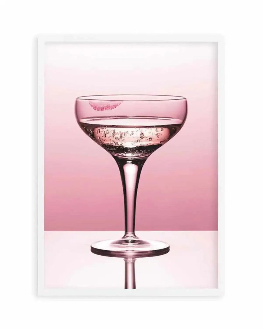 Framed photography art print of a coupe glass with a pink lipstick kiss print on the rim, displayed in a white frame with white border. The glass contains a pale pink sparkling beverage with visible bubbles, set against a two-toned pink background and reflected on a light pink surface below.