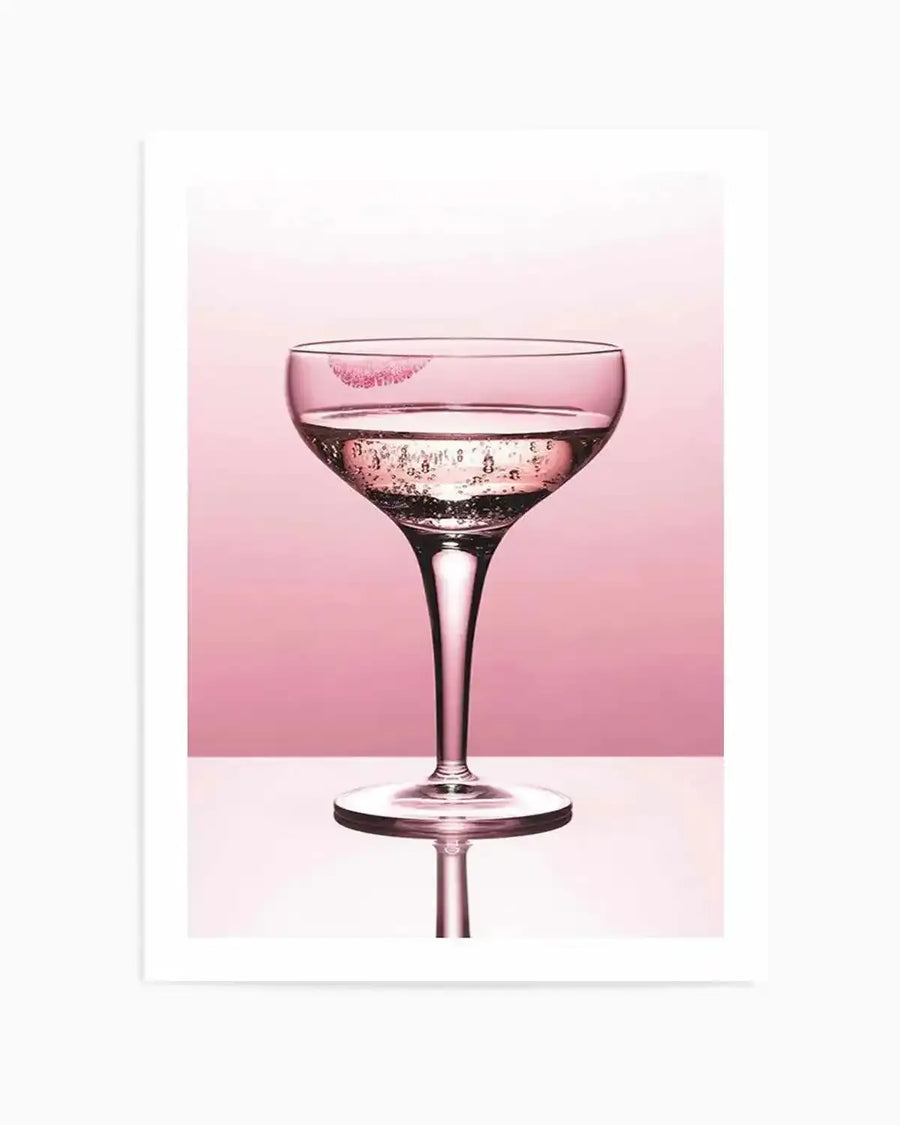 "Photography art print featuring a coupe glass filled with sparkling pink liquid, with a bright pink lipstick kiss mark on the rim, set against a two-toned pink background. The glass is reflected on a glossy white surface below."