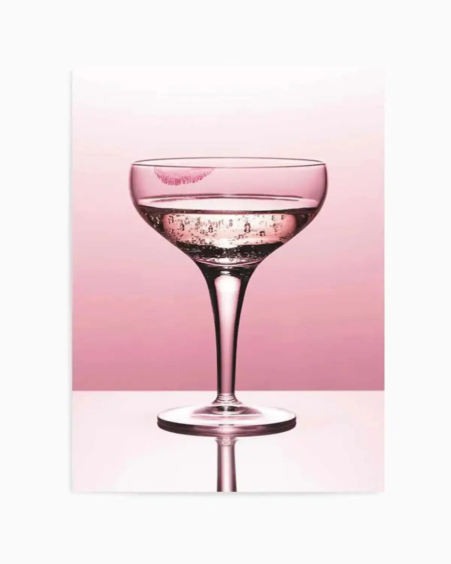 "Photography art print featuring a coupe glass filled with sparkling champagne, with a bright pink lipstick kiss mark on the rim, set against a soft pink background. The glass reflects on a white surface below, creating a symmetrical reflection of its stem and base."